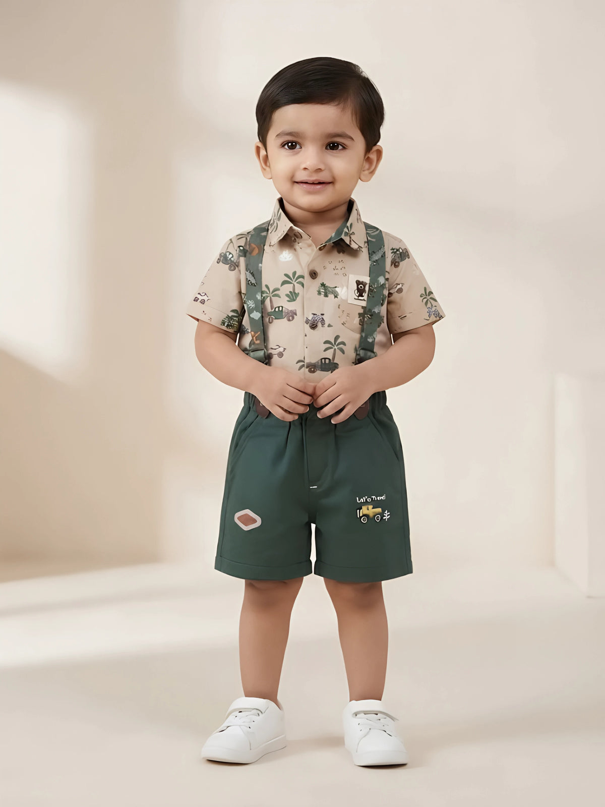 Infant boy wearing a beige printed short-sleeve shirt with green dungaree suspender shorts.