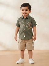 Young boy wearing an olive green short-sleeve printed shirt with a collar and beige cargo shorts with side flap pockets.