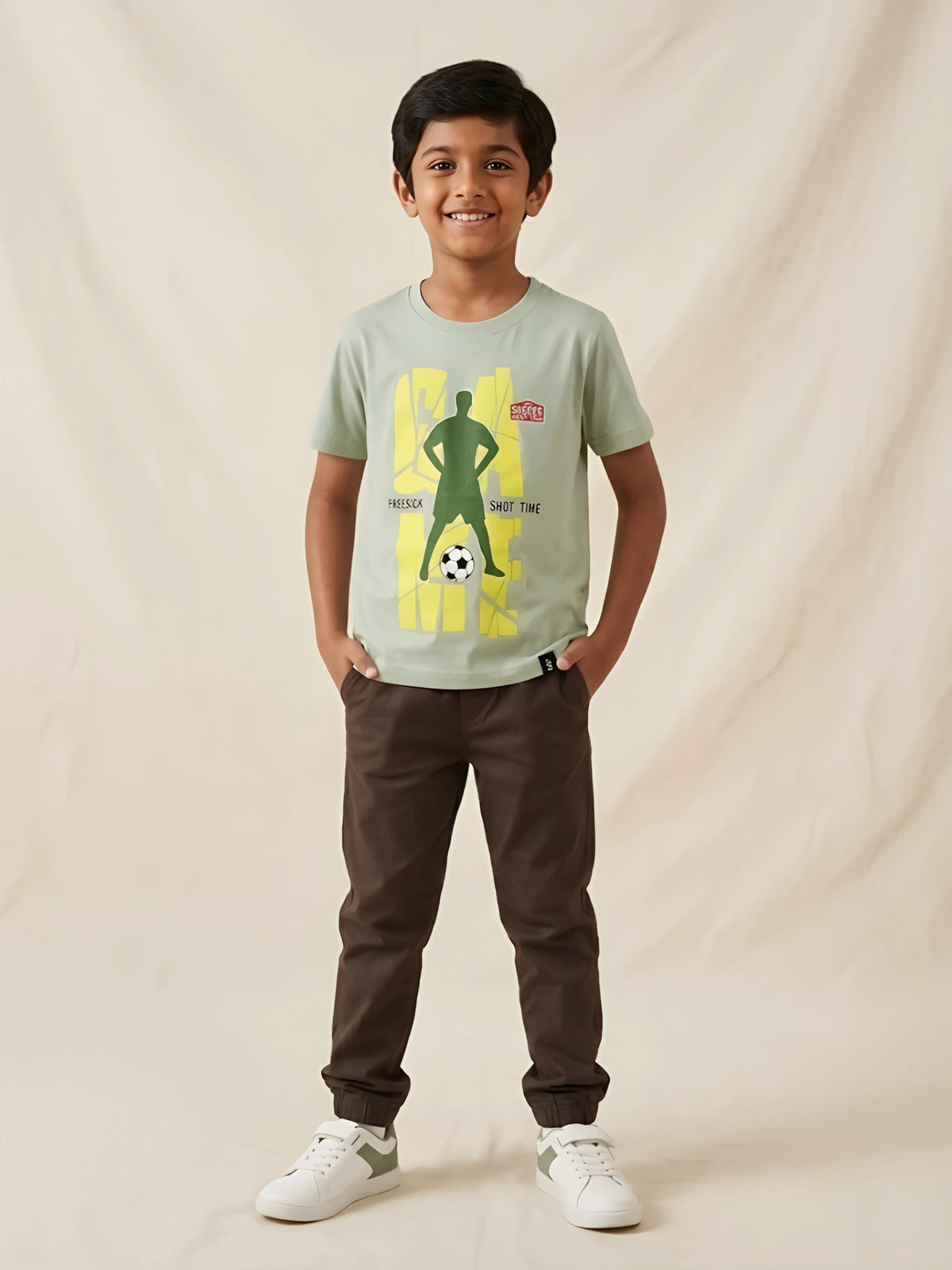 Young boy wearing a sage green short sleeve t-shirt featuring a football player graphic print, paired with brown trousers.
