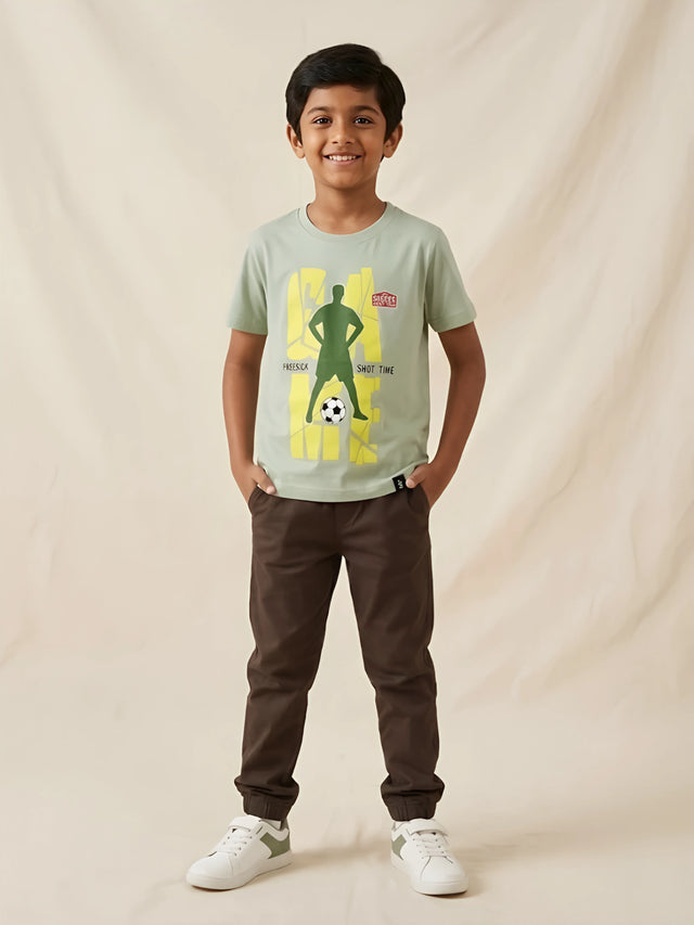 Young boy wearing a sage green short sleeve t-shirt featuring a football player graphic print, paired with brown trousers.