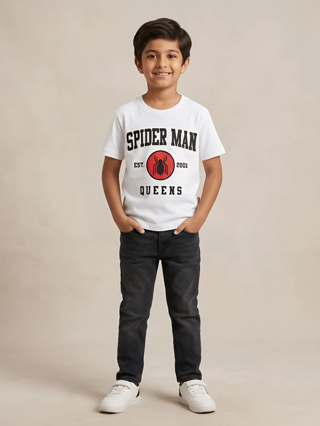 Young boy wearing a white short-sleeve graphic t-shirt with Spider-Man text and logo on the front paired with dark jeans.