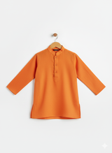 A young boy standing and smiling, wearing a bright orange long-sleeved kurta and pajama set, accessorized with a sleeveless teal, orange, and cream geometric-printed Nehru jacket.