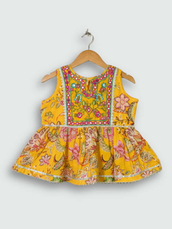 Front view of a girls' sleeveless yellow peplum top with all-over pink/green floral print and a heavily embroidered neckline and yoke, hanging on a wooden hanger.