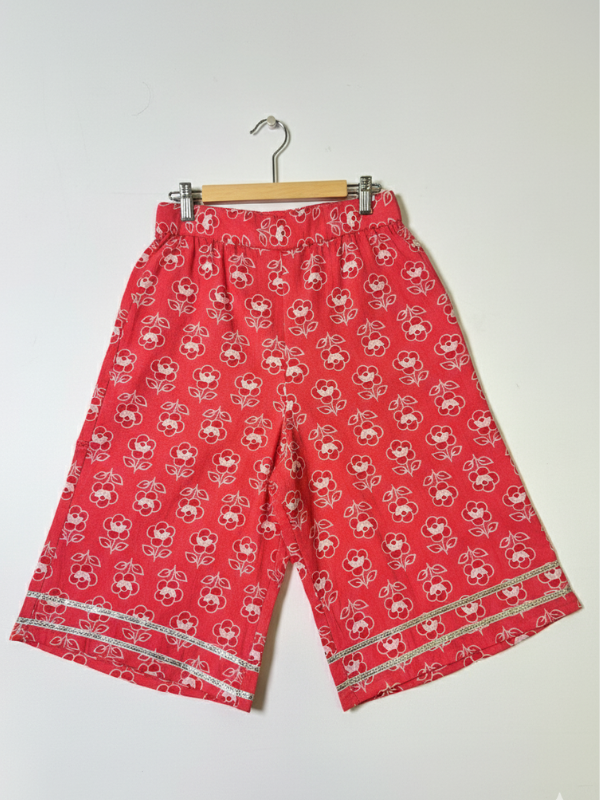 Full-length image of a young girl wearing a coral red and white printed peplum kurta with shoulder ties, wide-leg palazzo pants, and a white-to-red ombre dupatta with silver trim.