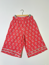 Full-length image of a young girl wearing a coral red and white printed peplum kurta with shoulder ties, wide-leg palazzo pants, and a white-to-red ombre dupatta with silver trim.