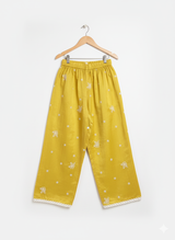 Front view of girls' wide-leg yellow palazzo pants with scattered white floral embroidery and a white lace trim on the hem, hanging on a wooden hanger.