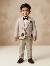 A young boy wearing a cream 3-piece coat suit with floral embroidery, matching trousers, a white formal shirt, and a black bow tie.