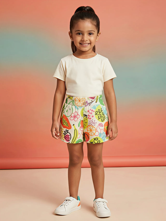 A young girl wearing bright yellow shorts with a colorful tropical fruit and doodle print, featuring an elasticated waistband and button detail.