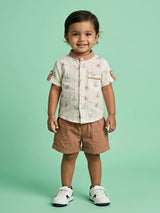 A young boy wearing a cream short-sleeve shirt with a sailboat print, mandarin collar, and front pocket, paired with matching solid brown shorts.