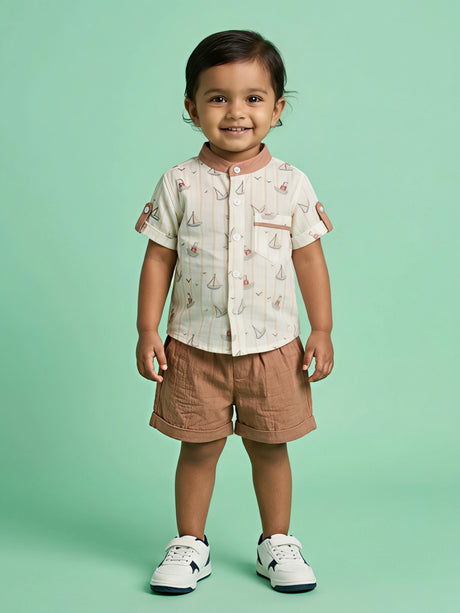 A young boy wearing a cream short-sleeve shirt with a sailboat print, mandarin collar, and front pocket, paired with matching solid brown shorts.