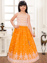 Young girl wearing a sleeveless orange and white lehenga choli set. The top is white with orange patterned embroidery, and the flared orange skirt features a thick white floral embroidered border.