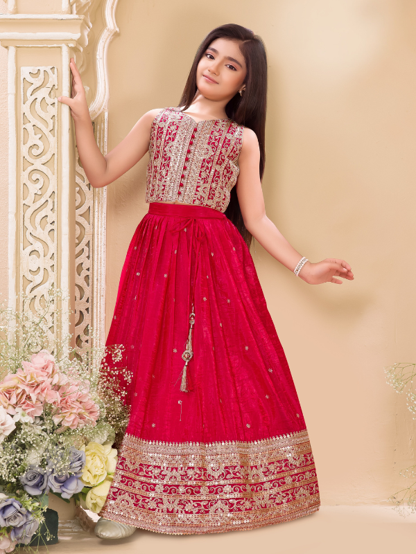 Young girl wearing a sleeveless deep red or magenta lehenga choli. The top is covered in gold and silver embroidery, and the skirt features a wide, heavily detailed gold zari and sequin border.