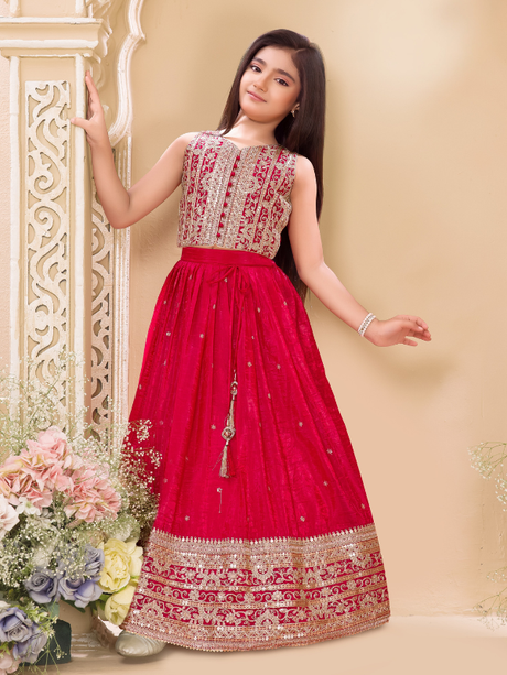 Young girl wearing a sleeveless deep red or magenta lehenga choli. The top is covered in gold and silver embroidery, and the skirt features a wide, heavily detailed gold zari and sequin border.