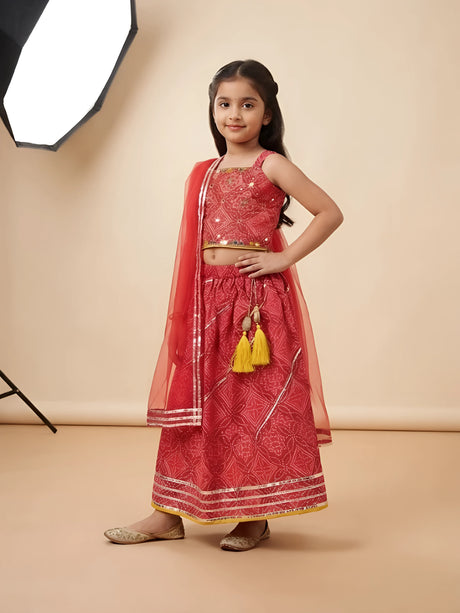 Young girl posing in a vibrant red printed lehenga choli set with gold gota patti border, yellow tassels, and sheer red dupatta