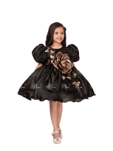 Young girl smiling in a black satin party dress with large puff sleeves and a metallic gold 3D rose accent on the bodice.
