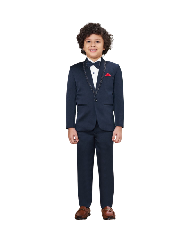 Young boy smiling and posing in a formal navy blue tuxedo suit set, featuring an embellished black lapel, a white shirt, a black bow tie, and a red pocket square. The look is completed with brown loafers