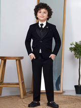Young boy with curly hair wearing a black textured coat suit with satin lapels, a black waistcoat, black trousers, a white shirt, and a black bow tie, standing indoors.