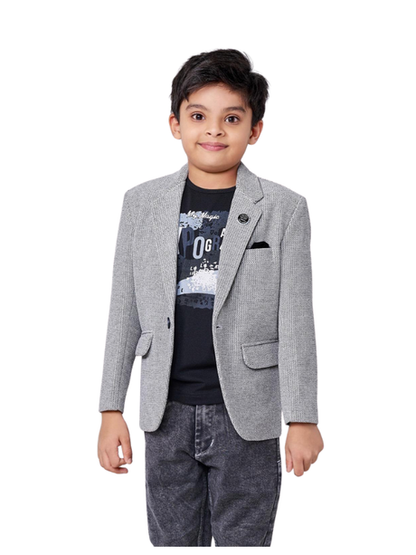Young boy smiling in a textured light grey blazer with a lapel pin and black pocket square, layered over a black graphic t-shirt and dark grey acid-wash jeans.