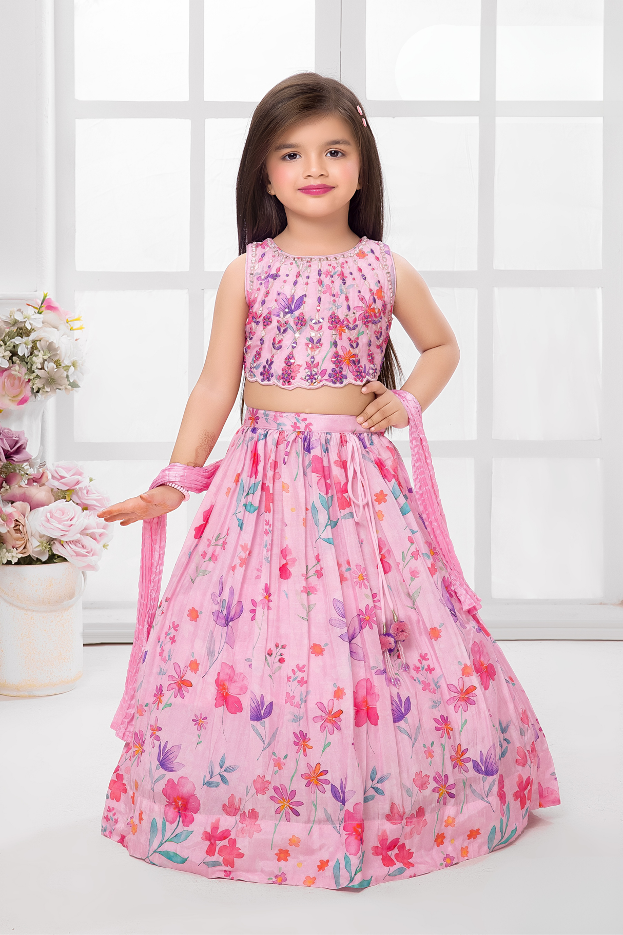 Young girl wearing a sleeveless light pink lehenga choli set with an all-over print of purple and pink flowers, featuring an embellished crop top.