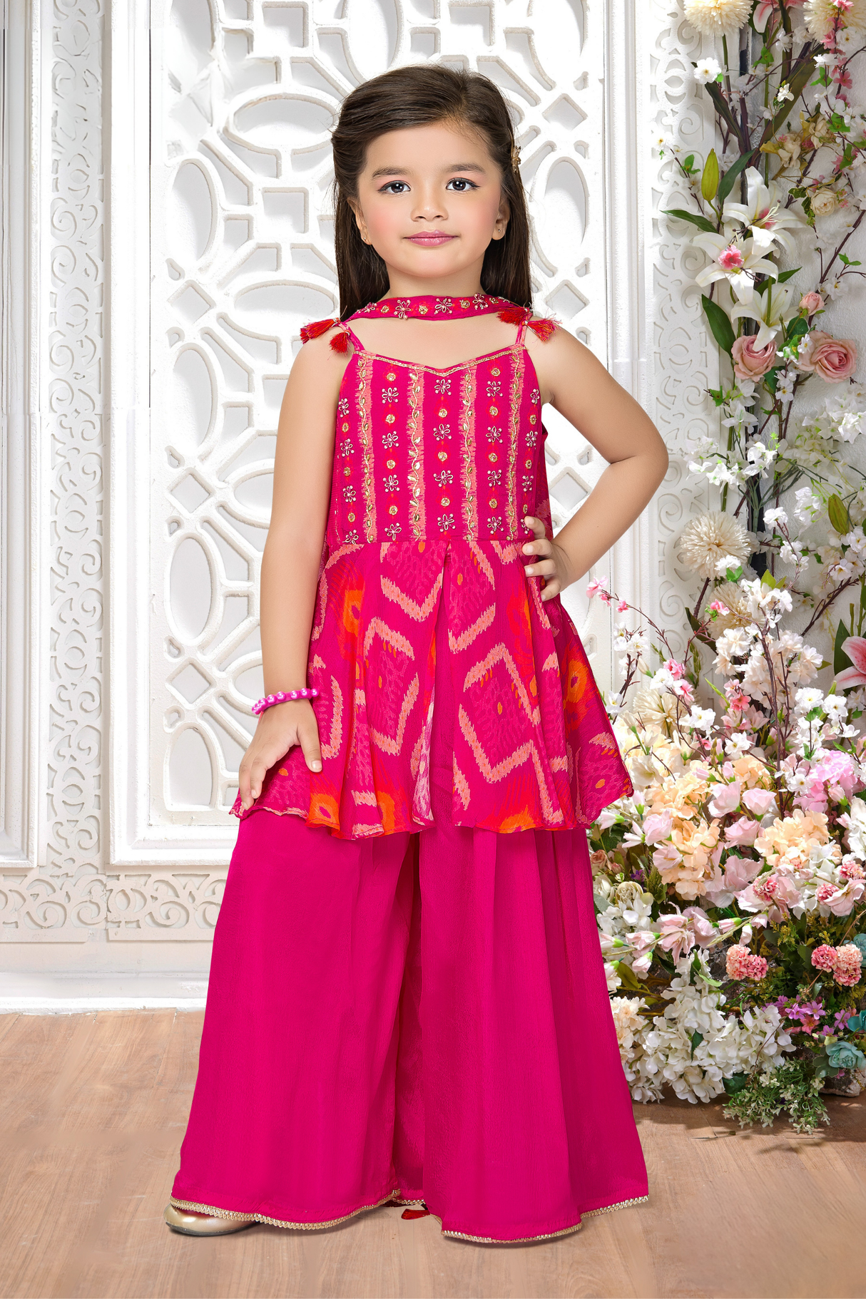 Young girl wearing a sleeveless Rani Pink sharara suit with a peplum top embroidered in gold thread and mirror work, paired with flowing matching sharara pants.