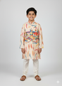A young boy standing and smiling, wearing a cream long-sleeved kurta and pajama set with a sleeveless, brightly colored abstract-printed Nehru jacket.