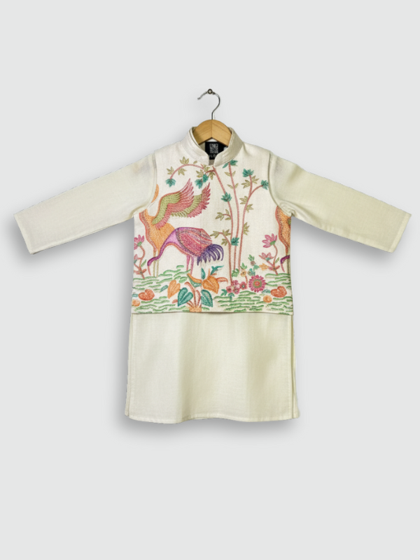 A young boy standing and smiling, wearing a cream/off-white long-sleeved kurta and pajama set with beautiful, detailed multi-color bird and floral embroidery across the chest.