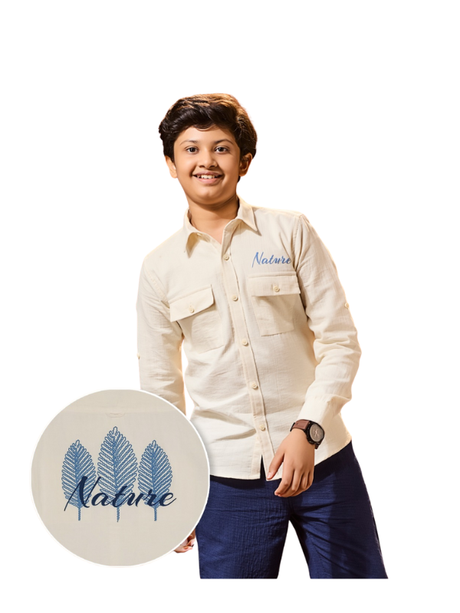 Young boy smiling and posing in a cream/light beige long-sleeve shirt with double flap chest pockets and blue 'Nature' embroidery, paired with dark blue trousers and a watch.