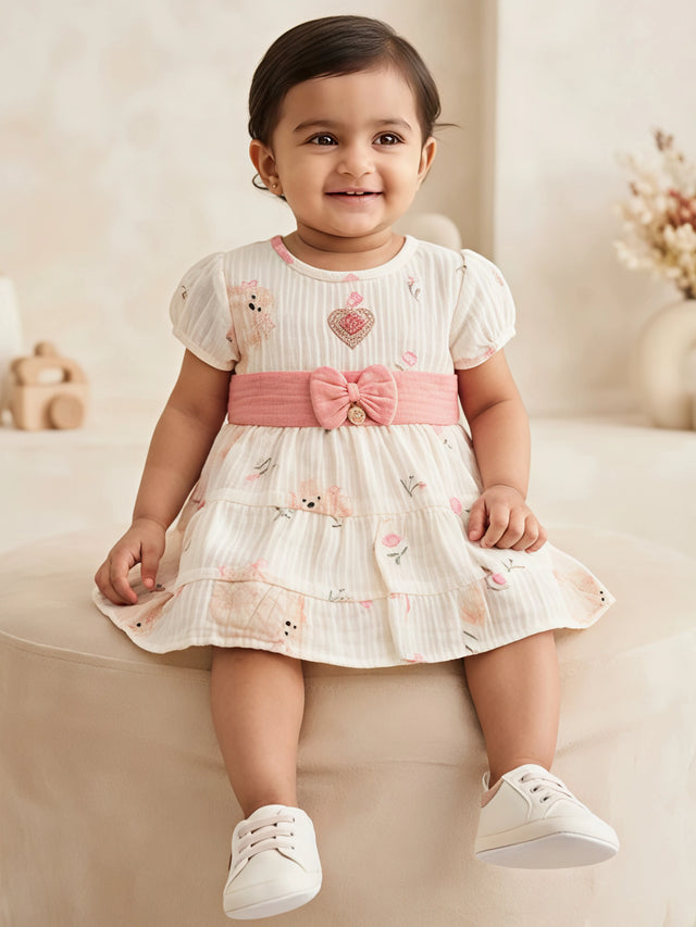 Front view of a baby girl wearing a cream printed frock with short puff sleeves, round neckline, pink bow waist band, and tiered skirt.