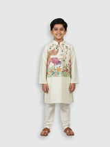 A young boy standing and smiling, wearing a cream/off-white long-sleeved kurta and pajama set with beautiful, detailed multi-color bird and floral embroidery across the chest.