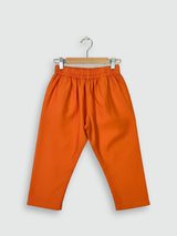 A young boy standing and smiling, wearing a bright orange long-sleeved kurta and pajama set, accessorized with a sleeveless teal, orange, and cream geometric-printed Nehru jacket.