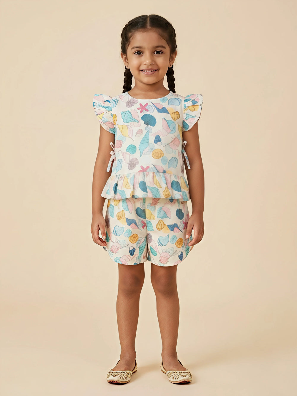 A young girl wearing a matching mint green and off-white shorts set featuring a colorful sea shell print, a peplum top with flutter sleeves, and side ties.