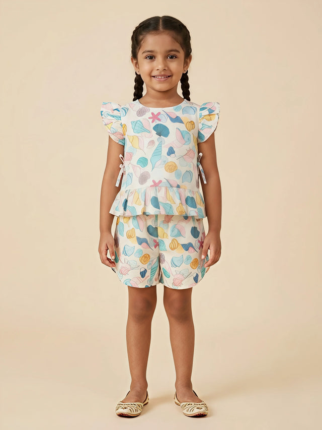 A young girl wearing a matching mint green and off-white shorts set featuring a colorful sea shell print, a peplum top with flutter sleeves, and side ties.