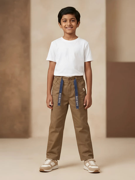 Young boy wearing brown casual trousers with navy blue printed tape detail and a white t-shirt.