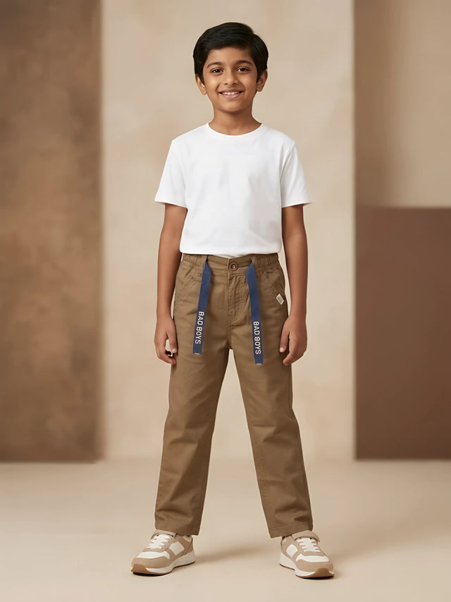 Young boy wearing brown casual trousers with navy blue printed tape detail and a white t-shirt.