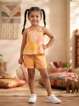 Young girl smiling in a yellow and pink tropical floral print shorts and top set, holding a straw hat.