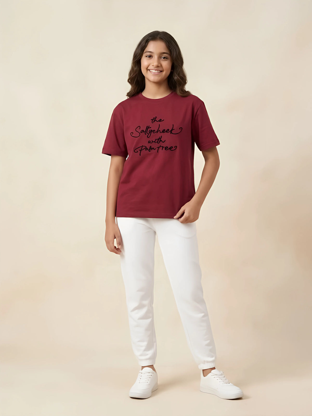 Young girl wearing a deep red short-sleeve casual t-shirt with black cursive text print, paired with white pants and sneakers.