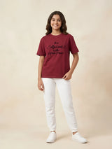 Young girl wearing a deep red short-sleeve casual t-shirt with black cursive text print, paired with white pants and sneakers.
