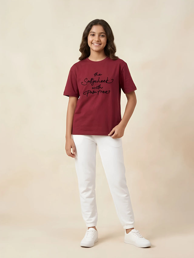 Young girl wearing a deep red short-sleeve casual t-shirt with black cursive text print, paired with white pants and sneakers.