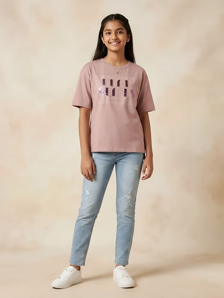 Young girl wearing a peach graphic casual top with half sleeves and light-wash blue jeans