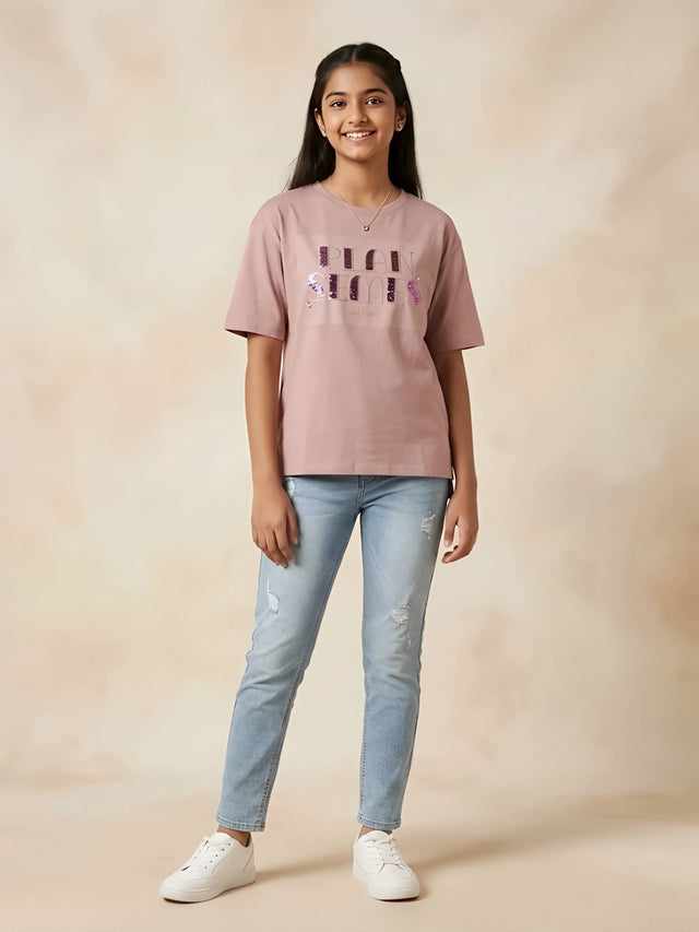 Young girl wearing a peach graphic casual top with half sleeves and light-wash blue jeans
