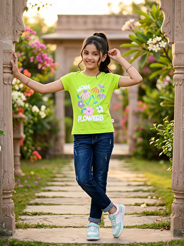 A young girl smiling in a lime green graphic t-shirt with a colorful floral print that reads "MORE FLOWERS", paired with blue jeans and sneakers.
