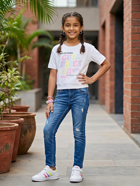 Young girl smiling and posing in a stylish white short-sleeve top with multi-colored pastel graphic text on the front.