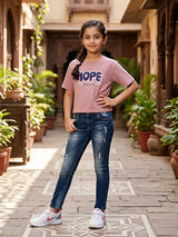 Young girl posing in a courtyard wearing a mauve purple casual top with 'HOPE' graphic print, paired with blue jeans and white sneakers.