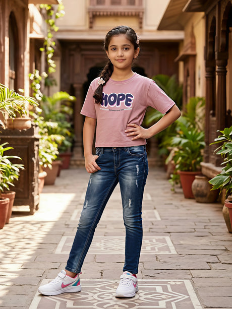 Young girl posing in a courtyard wearing a mauve purple casual top with 'HOPE' graphic print, paired with blue jeans and white sneakers.