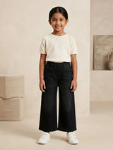 Young girl wearing black wide-leg casual pants with an elastic waistband and front pockets paired with a cream top and white sneakers.