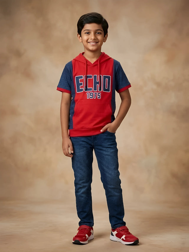 Young boy wearing a red and navy blue hooded short sleeve graphic t-shirt with kangaroo pocket paired with dark blue jeans