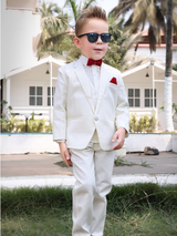 Full-length photo of a stylish young boy wearing a crisp white suit (jacket, vest, pants), black sunglasses, and a bright red bow tie and pocket square, posing outdoors in front of white cabanas.