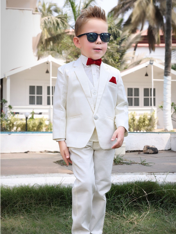 Full-length photo of a stylish young boy wearing a crisp white suit (jacket, vest, pants), black sunglasses, and a bright red bow tie and pocket square, posing outdoors in front of white cabanas.