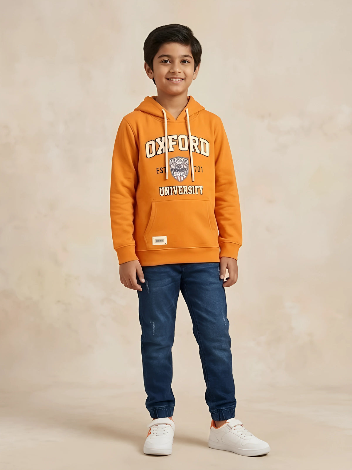Young boy wearing an orange long-sleeve hooded t-shirt with Oxford University graphic print and kangaroo pocket paired with blue jeans.