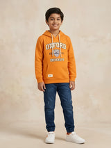Young boy wearing an orange long-sleeve hooded t-shirt with Oxford University graphic print and kangaroo pocket paired with blue jeans.